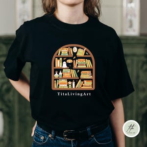 May include: Black t-shirt featuring a whimsical bookshelf design filled with books, a ghost, and other decorative elements. The text "TitaLivingArt" is printed below the design. The shirt is made of a soft material.