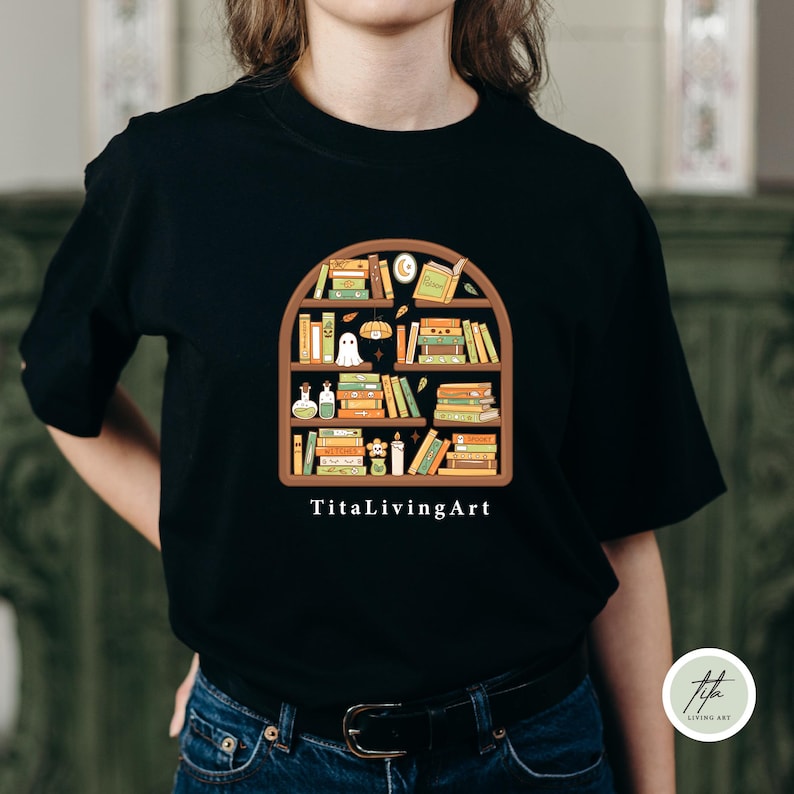 May include: Black t-shirt featuring a whimsical bookshelf design filled with books, a ghost, and other decorative elements. The text "TitaLivingArt" is printed below the design. The shirt is made of a soft material.