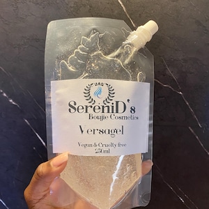 Peut inclure: Une poche en plastique transparente remplie d'un gel chatoyant de couleur claire. La poche est munie d'une étiquette blanche avec le texte "SereniD's Boujie Cosmetics Versagel Vegan & Cruelty free 250ml".