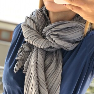 Op de afbeelding: Een grijze geplooide sjaal geknoopt om de nek van een persoon die een blauw shirt en jeans draagt.