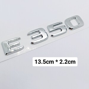 Peut inclure: Emblème E350 chromé, d'environ 13,5 cm sur 2,2 cm, sur fond blanc. Les chiffres sont en gras, de style sans-serif. Les dimensions sont affichées dans l'image.