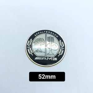 Könnte beinhalten: Ein rundes Emblem in Schwarz und Silber mit dem Text "Affalterbach" und "AMG" in silberner Schrift. Das Emblem zeigt einen Baum und einen stilisierten Motor-Kolben. Das Emblem hat einen Durchmesser von 52 mm.