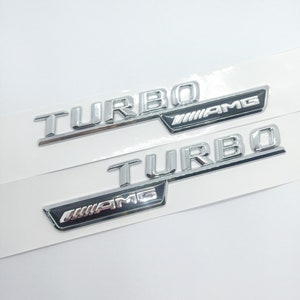 Könnte beinhalten: Zwei silberne Autoembleme mit dem Wort "TURBO" und dem Logo "AMG" in Schwarz.