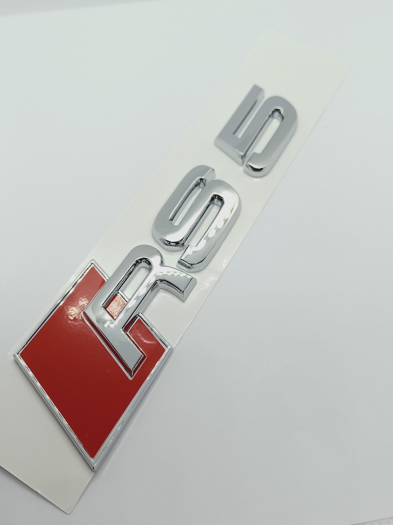 K&ouml;nnte beinhalten: Ein silbernes und rotes Autoemblem mit den Buchstaben "RS5" in erhabener Schrift.