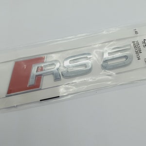 K&ouml;nnte beinhalten: Ein silbernes "RS 5" Emblem mit rotem Hintergrund. Das Emblem ist in einer durchsichtigen Plastikt&uuml;te verpackt.