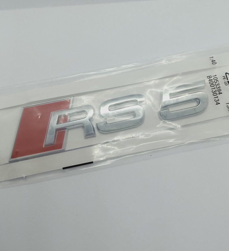 K&ouml;nnte beinhalten: Ein silbernes "RS 5" Emblem mit rotem Hintergrund. Das Emblem ist in einer durchsichtigen Plastikt&uuml;te verpackt.