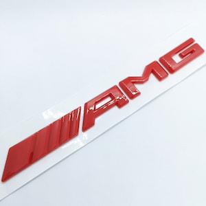 Könnte beinhalten: Rotes 3D-Plastik-Emblem "AMG" mit rotem Streifenmuster. Das Emblem befindet sich auf einem weißen Hintergrund.