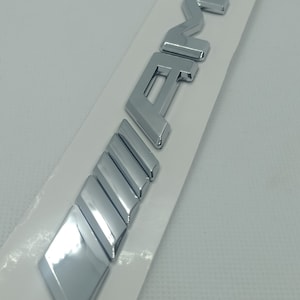 Może przedstawiać: Srebrny chromowany emblemat samochodowy z napisem "AMG" w wypukłych literach. Emblemat ma szereg pionowych linii pod napisem.