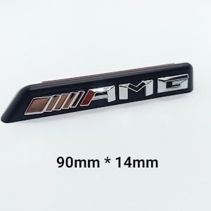 Puede incluir: Un emblema AMG negro y plateado, que mide 90 mm x 14 mm. El emblema presenta el logotipo AMG en letras plateadas y un diseño rojo y plateado. El emblema es rectangular con bordes redondeados.