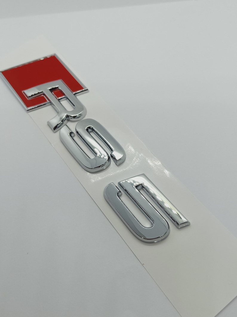 K&ouml;nnte beinhalten: Ein silbernes Chrom-Autoemblem mit den Buchstaben "RS5" in einer fetten, stilisierten Schrift. Das "R" ist rot.