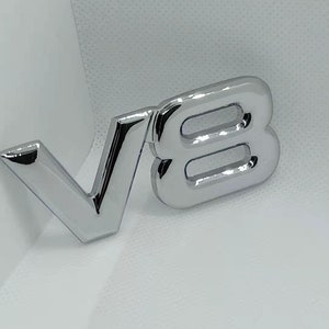 Peut inclure: Emblème en métal chromé avec les lettres "V8" dans une police audacieuse en trois dimensions.