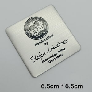 May include: A silver metal square plaque with a black and white logo and text. The logo is a circle with a tree and a building inside. The text reads "Handcrafted by Stefan Karcher Mercedes-AMG Germany". The plaque measures 6.5 cm by 6.5 cm.