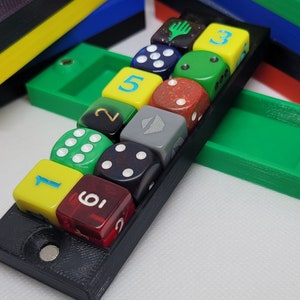 May include: A black dice tray holding twelve multi-colored dice. The dice are various colors, including red, green, yellow, and gray, with white or black pips. Several other colored trays are stacked in the background.