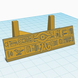 May include: A gold-colored stand with two upright supports and a horizontal platform. The platform features a raised design of an eye with hieroglyphics on either side.