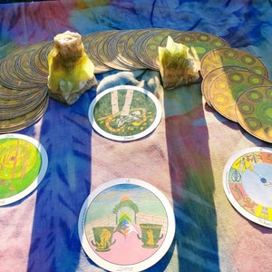 May include: A tarot card reading spread with four cards laid out on a colorful fabric background. The cards are "Death", "Wheel of Fortune", "Lovers", and "The Tower". Two yellow crystals are placed on either side of the spread.