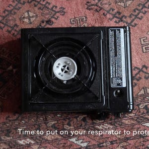 May include: A black portable gas stove with a silver burner is centered on a patterned red rug. The stove has a warning label on the side. A rope is in the upper right corner and a smaller rope is in the lower left corner.