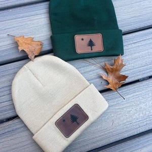 May include: Two knit beanies with a brown leather patch featuring the state of Maine flag. One beanie is green and the other is beige.