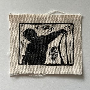 May include: Black and white linocut print of a person holding a long, thin object. The print is framed in a black square and is mounted on a white fabric background.