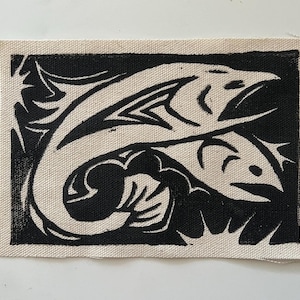 May include: A rectangular patch featuring a black and white linocut print of two fish leaping. The fish are depicted with stylized details against a black background, surrounded by abstract shapes. The patch has a beige border.