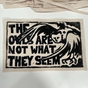 May include: A rectangular fabric patch featuring a black screen-printed graphic on a natural off-white canvas background. The design displays a stylized owl with wings spread and the text "THE OWLS ARE NOT WHAT THEY SEEM" in bold black lettering.