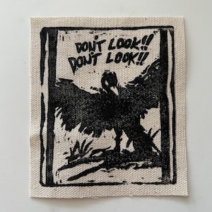 May include: A square, off-white fabric patch with a black illustration of a bird with outstretched wings. The text "DON'T LOOK!! DON'T LOOK!!" is printed above the bird in black.