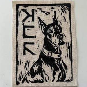 May include: A black and white linocut print of a Doberman Pinscher dog on a beige fabric patch. The dog is wearing a collar and the word "KUK" is printed vertically on the left side of the patch.