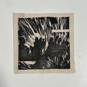 May include: A square, black and white linocut print on a beige fabric square. The print depicts a crowd of people and abstract shapes. The image has a handmade, artistic feel.