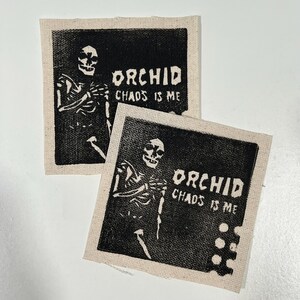 May include: Two square patches with a black and white skeleton graphic and the text "ORCHID CHAOS IS ME". The patches are printed on a beige fabric.