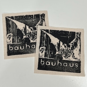 May include: Two square, off-white fabric patches with a black Bauhaus design. The design features an abstract architectural image and the word "bauhaus" in a sans-serif font. The patches are likely for sewing or crafting.