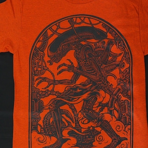 May include: Orange t-shirt featuring a black illustration of an alien creature within an arched frame. The design includes intricate mechanical details and a stylized depiction of the alien. The shirt is made of a soft material.