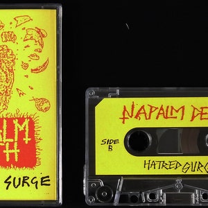 Op de afbeelding: Een gele cassetteband met de bandnaam "NAPALM DEATH" en de albumtitel "HATRED SURGE" in rood gedrukt. De band heeft een schedelgrafiek aan de voorkant.