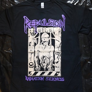Puede incluir: Camiseta negra con un gráfico blanco y morado de una figura esquelética en una jaula. El texto "Repulsion" y "Radiation Sickness" está impreso en morado.
