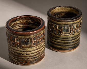 Peut inclure: Deux tasses cylindriques en céramique avec un design texturé et terreux. Les tasses présentent un mélange de tons bruns, beiges et verts, avec un motif carré répétitif sur la partie supérieure. Les bords sont brun foncé.