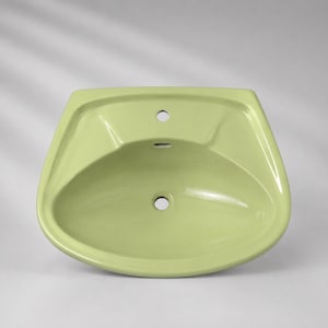 May include: A light sage green ceramic bathroom sink basin with a glossy finish. The basin features a central faucet hole at the top, a small overflow drain opening below it, and a larger circular drain hole located at the bottom centre.