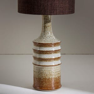 Puede incluir: Una lámpara de mesa de cerámica con una pantalla de tela marrón. La base de la lámpara es de color beige y marrón moteado con bandas horizontales. La lámpara tiene un cuello largo y delgado y una pantalla redonda.