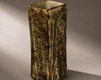 Puede incluir: Un jarrón de cerámica alto y rectangular con una superficie texturizada similar a la corteza. El exterior presenta un esmalte en tonos marrón oscuro, verde oliva y crema, creando un aspecto veteado vertical. La abertura superior es cuadrada.