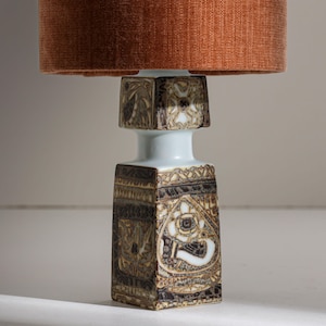 Puede incluir: Lámpara de mesa decorativa con base cuadrada y parte superior cilíndrica. La base presenta un diseño detallado con patrones en tonos marrón, negro y blanco. La pantalla es de color naranja quemado, añadiendo un toque vintage.