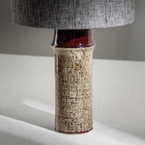 Puede incluir: Una lámpara de mesa de cerámica con una pantalla de tela gris. La base de la lámpara es de forma cilíndrica con un esmalte texturizado y moteado en tonos de marrón, verde y blanco.