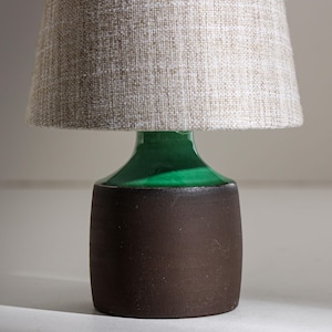 Puede incluir: Una pequeña lámpara de mesa con base marrón y cuello verde que sostiene una pantalla de tela beige. La lámpara tiene un diseño clásico y minimalista, adecuada para un dormitorio o sala de estar. Las dimensiones de la lámpara no son visibles.