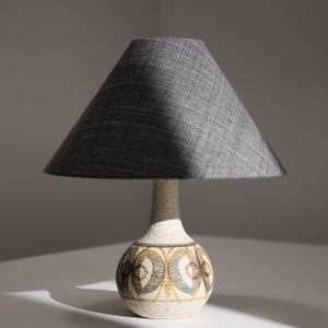 May include: A table lamp featuring a conical gray fabric lampshade and a textured, off-white ceramic base. The base has a rounded, bulbous shape decorated with a repeating pattern of brown and muted green circular motifs around the center.
