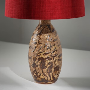 Puede incluir: Lámpara de mesa decorativa con pantalla de tela roja. La base de la lámpara es marrón con un diseño en relieve detallado con caballitos de mar y plantas acuáticas. La lámpara está diseñada para uso en interiores.