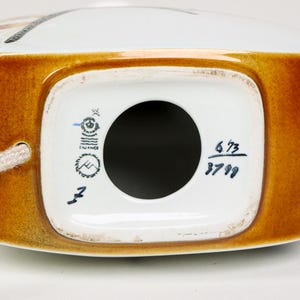 May include: The bottom of a white ceramic figurine with a brown glaze around the edges. The figurine has a blue and white mark with the text "Sweden" and a black circle with a mountain range inside. The bottom also has the numbers "6/73" and "3799" written in blue.
