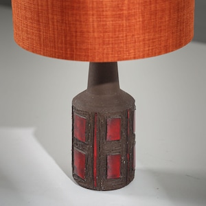 Puede incluir: Una lámpara de mesa moderna de mediados de siglo con una base de cerámica marrón texturizada con detalles rectangulares rojos. La lámpara tiene un cuello marrón oscuro y está rematada con una pantalla de tela naranja. La lámpara está diseñada para proporcionar iluminación ambiental.