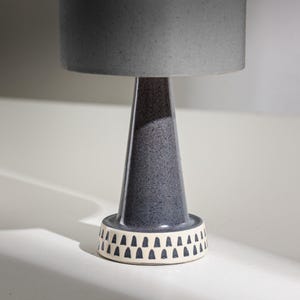 Puede incluir: Una lámpara de mesa moderna con una pantalla gris y una base de cerámica moteada azul oscuro. La base tiene una banda blanca con un patrón de triángulos negros repetidos. La lámpara tiene forma cónica.