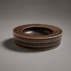 May include: A circular, low-profile ceramic bowl featuring a dark brown glaze with a speckled texture. The exterior surface is decorated with a series of horizontal, incised lines that wrap around the circumference of the vessel.