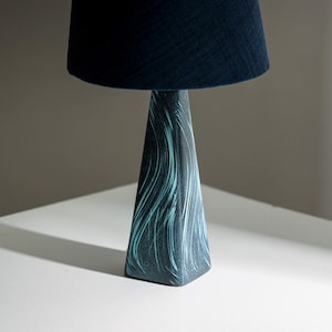 Puede incluir: Una lámpara de mesa de cerámica azul con una pantalla de tela azul oscuro. La base de la lámpara es una pirámide cuadrada con un patrón de espiral.