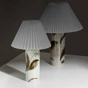 Könnte beinhalten: Zwei Keramik-Tischlampen mit gefalteten weißen Lampenschirmen. Die Lampen haben einen weißen Sockel mit braunen Blumendesigns.