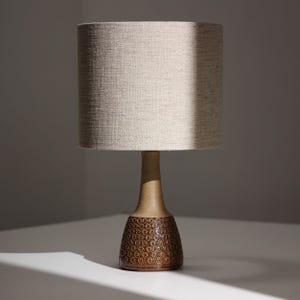 May include: A table lamp featuring a cylindrical beige fabric lampshade and a tapered ceramic base. The base has a smooth, light brown neck and a wider, dark brown bottom section decorated with a repeating pattern of small, embossed circular rings.