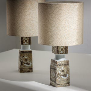 Puede incluir: Dos lámparas de mesa a juego. Cada lámpara tiene una pantalla beige con un estampado de hojas. La base tiene forma rectangular con un diseño decorativo blanco y marrón. Las lámparas están sobre una superficie blanca.