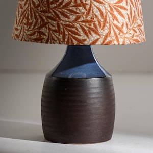 Puede incluir: Una lámpara de mesa de cerámica con base azul oscuro y pantalla con estampado botánico en tonos marrón y crema. La lámpara tiene base cilíndrica y parte superior acampanada.